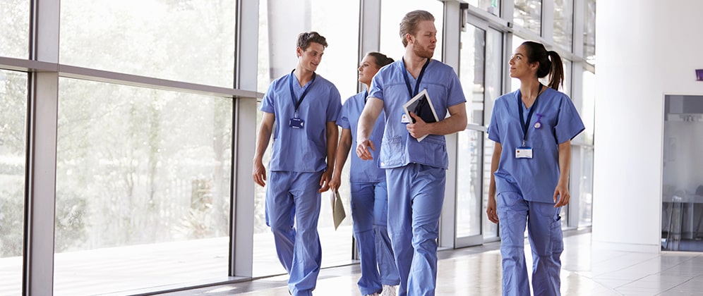 Four healthcare workers in scrubs walking in medical facility corridor talking to one another.