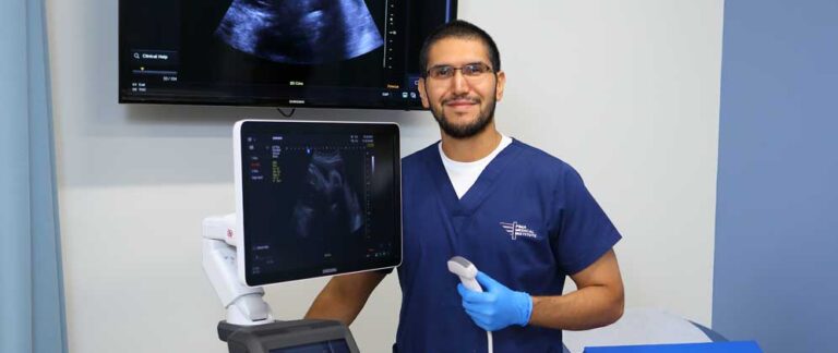 Man facing camera wearing blue Pima Medical scrubs standing with a diagnostic medical sonography machine