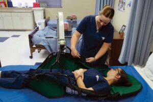 Image of a nursing assistant student helping another student into a lift during hands-on training in the lab.