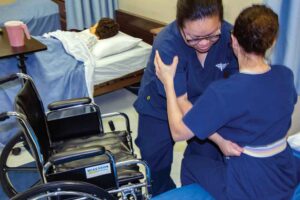 Image of a nursing assistant student helping another student into a wheelchair during hands-on training in the lab.