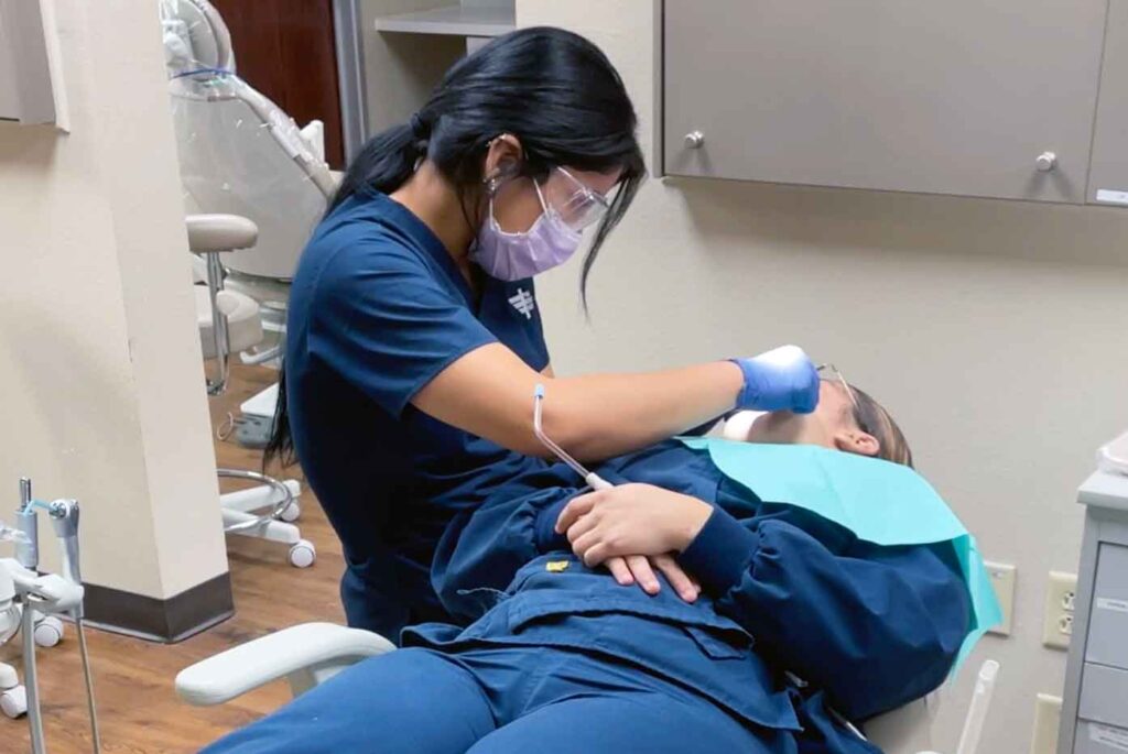 dental student in scrubs and PPE practices dental skills on classmate in lab classroom