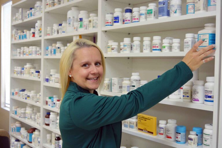 graduate smiling at camera while reaching toward shelving of prescriptions.