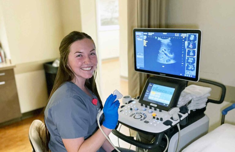 Diagnostic Medical Sonography student in lab setting sitting at ultrasound machine smiling.