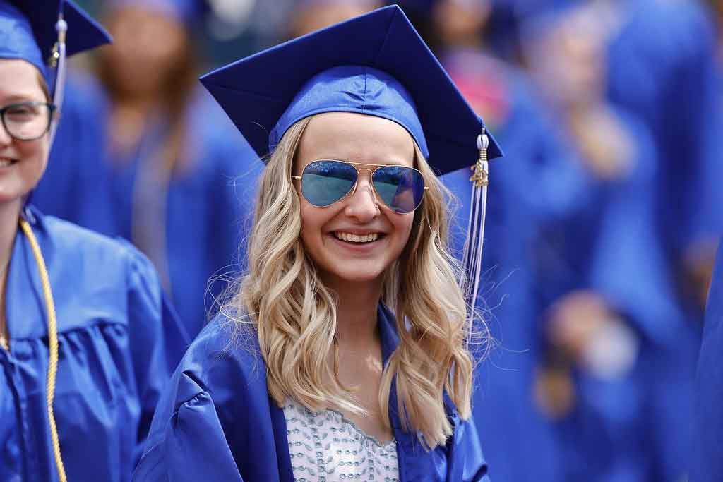 Student smiling at the camera in blue cap and gown and wearing sunglasses.