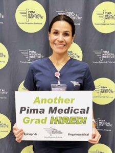 Phlebotomy graduate posing with a sign that says “Another Pima Medical Grad Hired”.