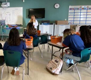 Students sit in desks facing away from camera and toward front of classroom as teacher instructs