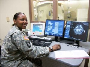 female soldier in Army uniform sitting at desk with radiologic images on computer screens in front of her.