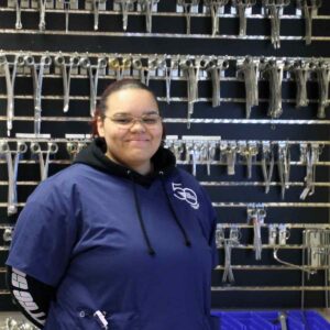 Younger woman in scrubs posing for camera in front of wall of tools used in medical procedures.