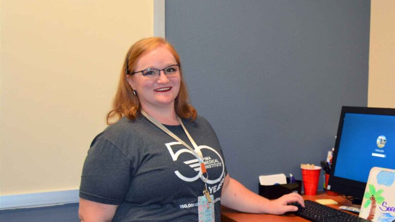 woman wearing Pima Medical t-shirt standing in front of computer monitor while smiling at the camera