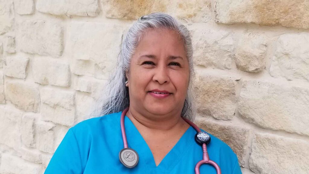 photo of woman leaning against a wall, wearing blue scrubs while facing the camera