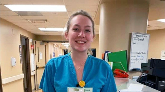 woman in light blue scrubs standing at hospital front desk holding up a piece of paper to the camera