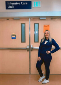 woman in all black scrubs posing for the camera in front of the door to the Hospital ICU department