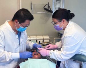 female in PPE gear and white lab coat seated across from male dentist in PPE and white lab coat as they perform a dental exam on a patient.