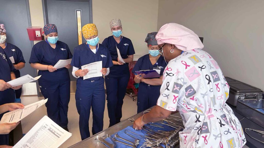 instructor in scrubs, hair covering, mask and gloves explains various surgical instruments to class of students, also wearing full PPE gear.