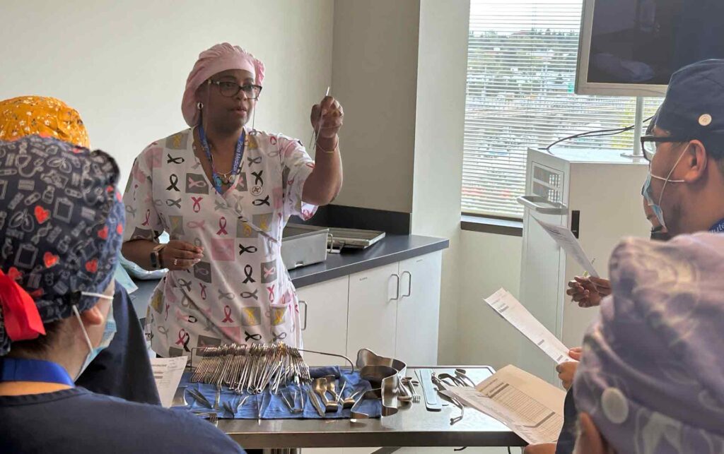 instructor dressed in PPE gear holds up surgical instrument for students to see.