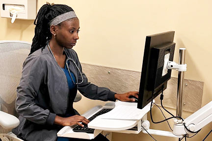 African-American Woman in scrubs with stethoscope around her neck sitting at a computer desk.