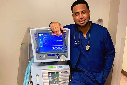 African- American man in scrubs facing the camera while leaning up against a piece of medical equipment.