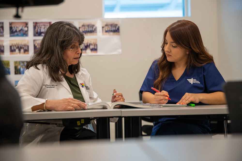 wide shot of instructor on the left with a female student in scrubs on the right. They are sitting at a table looking at a book together