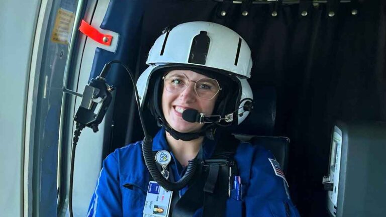 woman sitting in helicopter wearing flight gear while smiling at the camera.