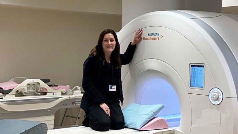 Female graduate kneels just outside a piece of radiology equipment while smiling at the camera.