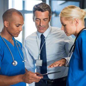 gray-haired man in dress shirt and tie discussing something with a male and female in blue scrubs.