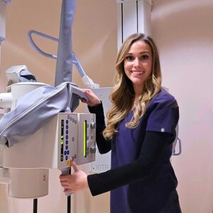 woman in black scrubs facing the camera while standing with medical imaging equipment.