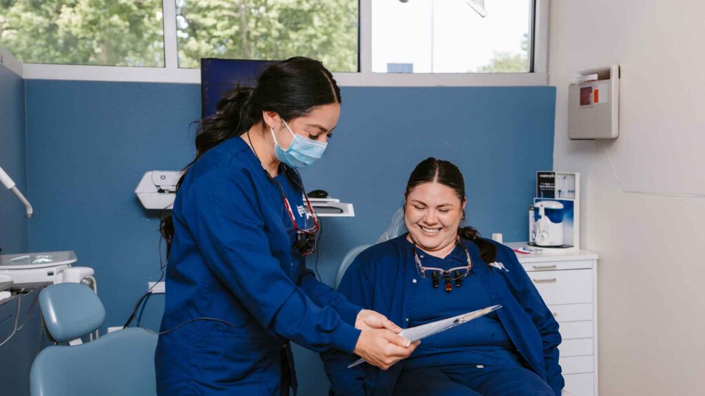 student in dental PPE reviews paperwork with another student who is portraying the role of patient sitting in the dental chair