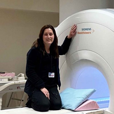 Korrine_H-hero Female graduate kneels just outside a piece of radiology equipment while smiling at the camera.